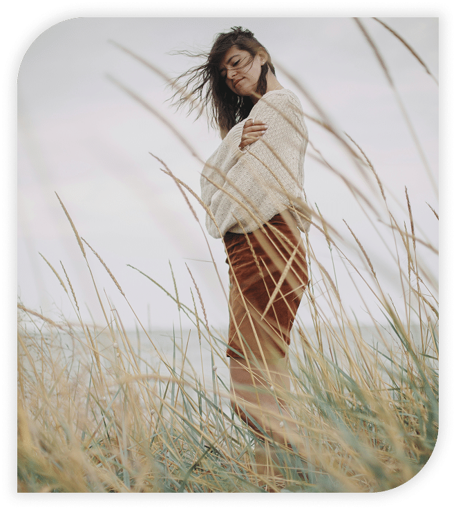 Woman standing in tall grass by the coast on a windy day, wearing a cozy sweater.
