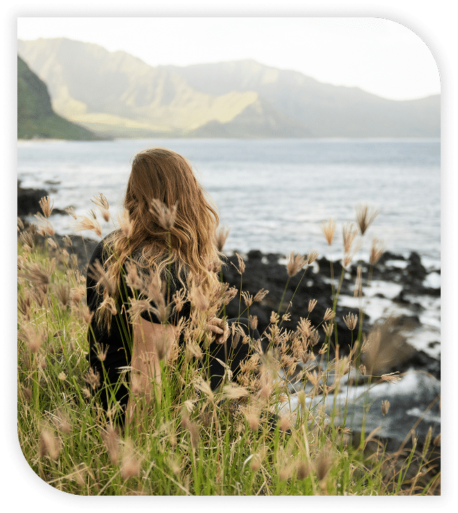Woman with long hair sitting in tall grass, overlooking the ocean and distant mountains.