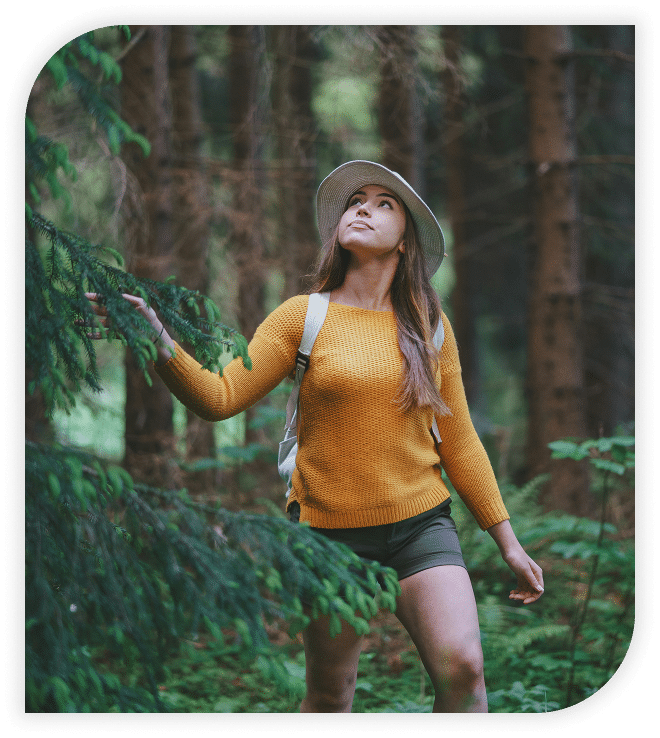 Woman in a yellow sweater and hat hiking through a forest, looking up at the trees.