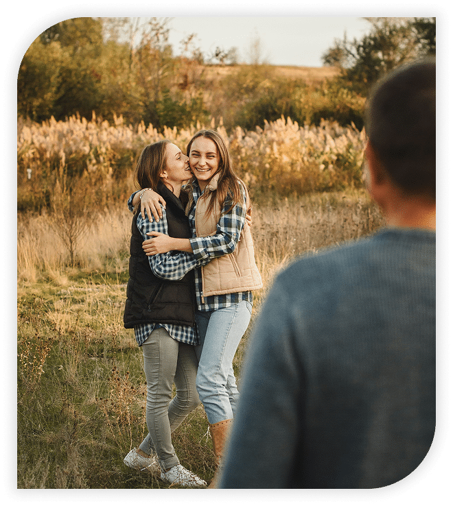 Couple hugging and smiling in a field as another person looks on.